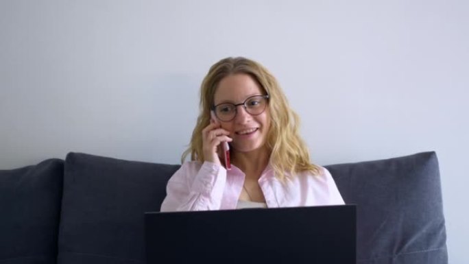 美丽的金发女人正坐在家里的笔记本电脑前，微笑着打电话。在家远程工作，不用去办公室。简化工作，降低成本