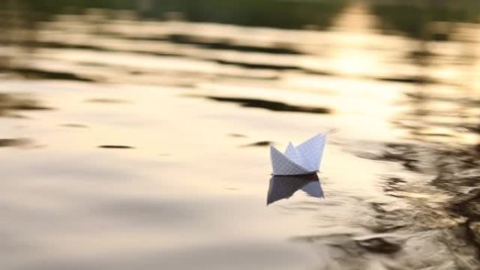 一艘纸船在美丽的日落时漂浮在水中的波浪上。折纸船航行。梦想，未来，童年，自由或希望的概念。慢动作。高