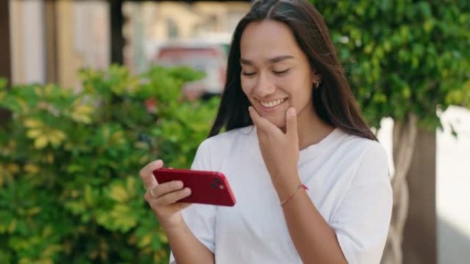 年轻美丽的西班牙裔妇女微笑着自信地在街上的智能手机上观看