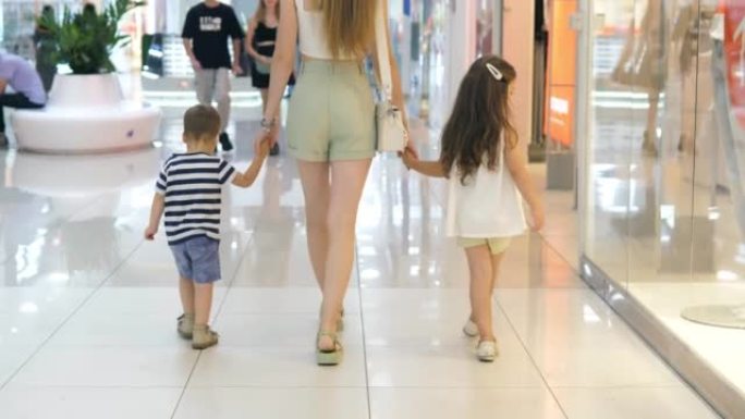 一个穿着时髦服装的年轻迷人女孩正带着她的孩子们在购物中心散步。妈妈和女儿和儿子牵着手去购物。一家人正
