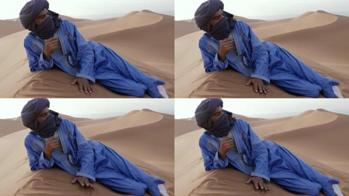 一名摩洛哥男子穿着蓝色的gandoura，djellaba和头巾，躺在摩洛哥Chigaga沙漠的沙丘