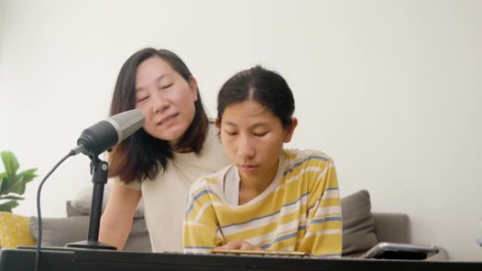 快乐的亚洲妈妈和正在玩电钢琴的女儿一起在沙发上唱歌，一起坐在家里的沙发上，生活理念。