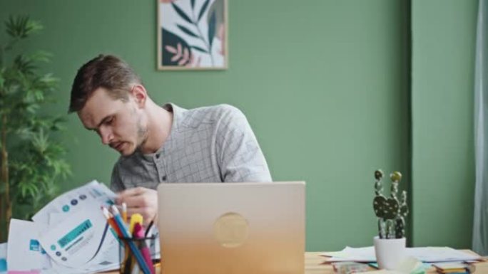 年轻的黑发男人坐在办公室里，有胡茬，感到压力很大，在笔记本电脑特写镜头的桌子上找不到合适的工作文件