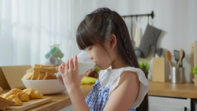 亚洲小孩拿着一杯牛奶，在家里的厨房里喝酒。年轻的学龄前女孩或女儿喝着牛奶，醒来后在家里接受保健后，在