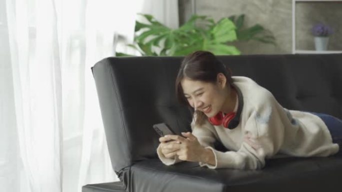 可爱的亚洲女人坐在沙发上用智能手机玩游戏。