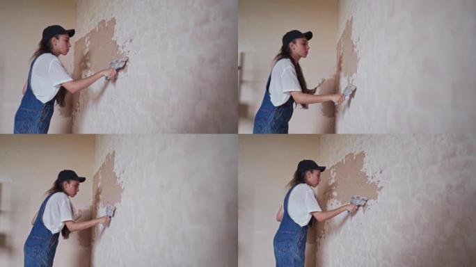 专业抹灰师女性在墙壁上撒上，在工作服和球帽的抹刀上涂抹并涂抹石膏。年轻女子膏墙，在建筑工地做装饰工作