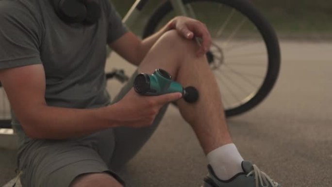 骑自行车的人在骑自行车锻炼后用按摩敲击器按摩肌肉和肌腱。运动员用手中的电动手枪按摩器按摩肌肉。运动恢