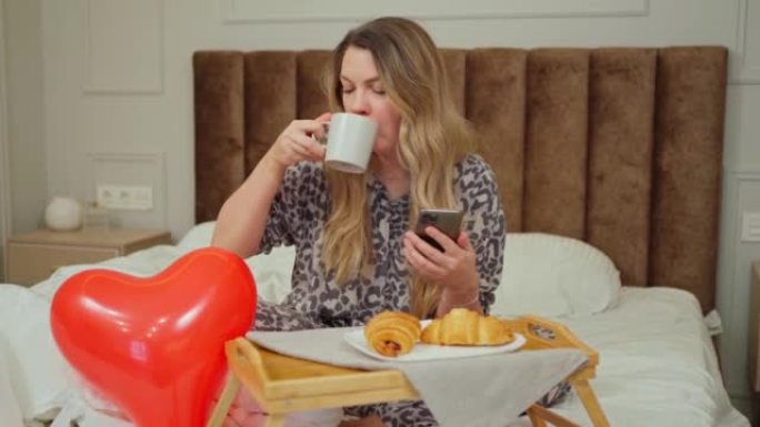一名年轻女子坐在床上，在床头柜上吃早餐，看着智能手机里的东西微笑着，气球旁边是一颗心。浪漫早餐