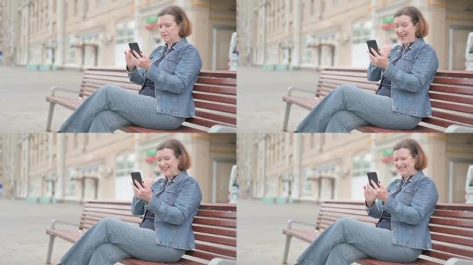 老妇人坐在户外长凳上庆祝智能手机在线成功