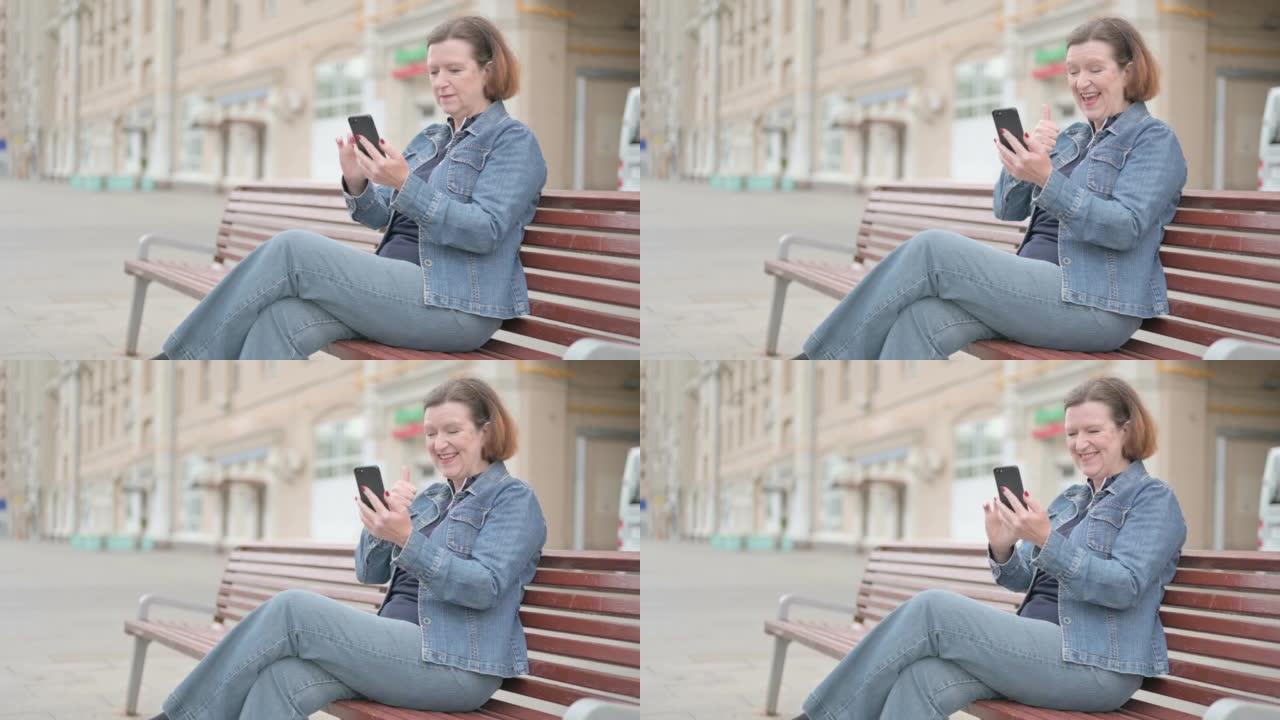 老妇人坐在户外长凳上庆祝智能手机在线成功