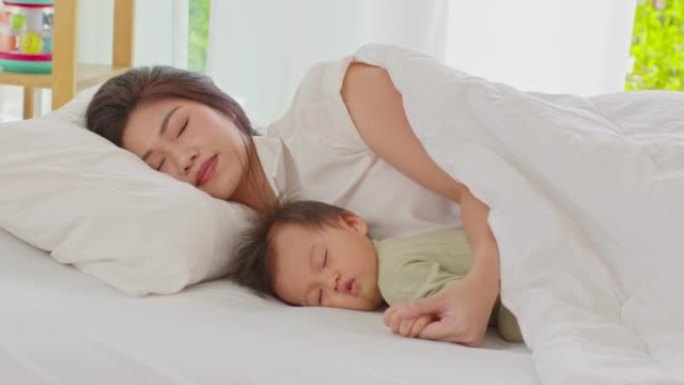 亚洲妈妈抱着婴儿的手躺在床上，一起爱睡。亚洲妈妈抱着她的婴儿放松，在温暖的地方躺在舒适的床上休息。婴