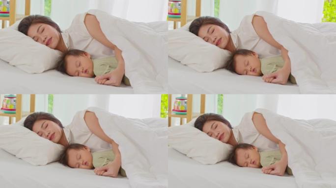 亚洲妈妈抱着婴儿的手躺在床上，一起爱睡。亚洲妈妈抱着她的婴儿放松，在温暖的地方躺在舒适的床上休息。婴