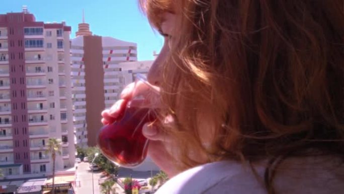 红头发的女人在阳光明媚的日子里在阳台上喝土耳其茶，向街上望去，风吹着头发
