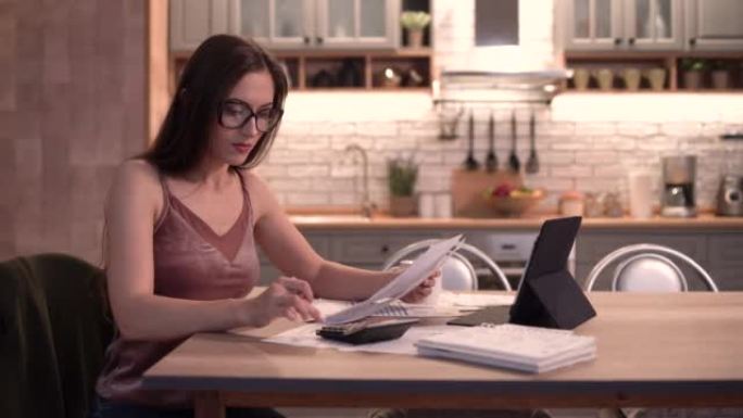 4 k视频，年轻女性晚上坐在厨房的桌子上，计算公用事业付款，在平板电脑上的在线银行应用程序中进行汇款