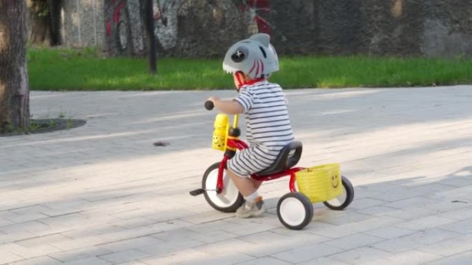 母亲教小孩在户外骑三轮车，蹒跚学步的男孩穿着有趣的自行车头盔在夏季公园玩得开心。儿童户外游乐场骑行活
