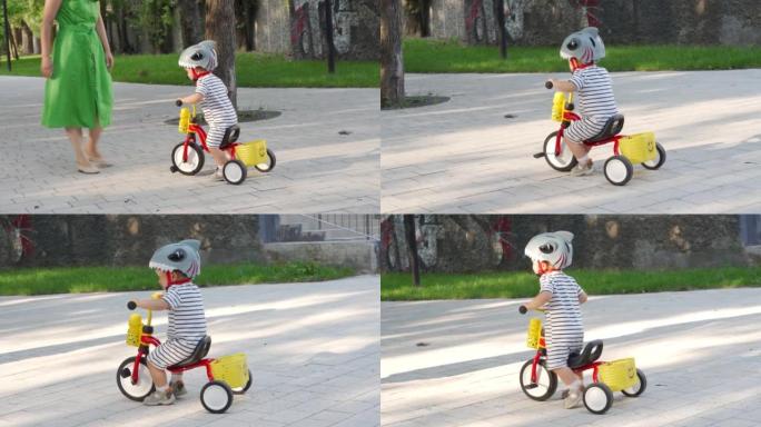 母亲教小孩在户外骑三轮车，蹒跚学步的男孩穿着有趣的自行车头盔在夏季公园玩得开心。儿童户外游乐场骑行活