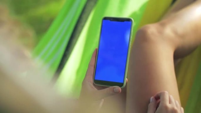女人躺在吊床上，用手机搜索互联网。戴着眼镜的年轻卷发女孩拿着智能手机，在公园里的树林中闲逛。蓝屏电话