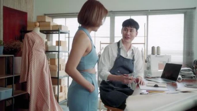 年轻的亚洲企业主男子与顾客女性讨论服装设计，穿着围裙的聪明男性接受客户订购新的系列服装。启动企业家f