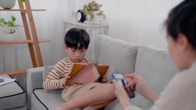4K，亚洲男孩坐在沙发上用平板电脑玩游戏，他的单身母亲坐在他旁边，用手机与朋友聊天。一中年男女坐在客