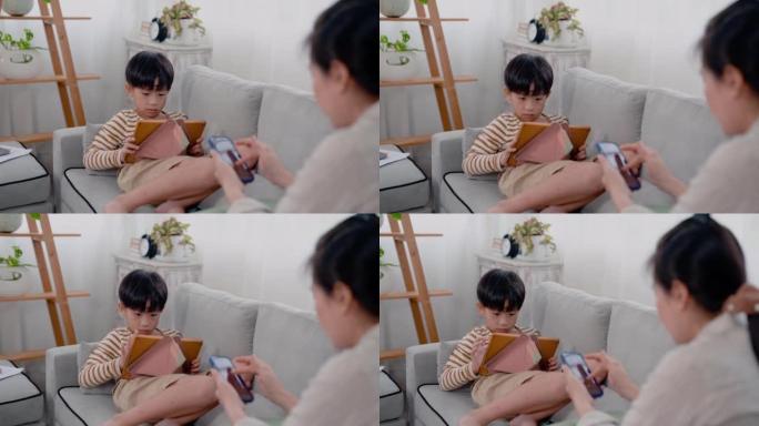 4K，亚洲男孩坐在沙发上用平板电脑玩游戏，他的单身母亲坐在他旁边，用手机与朋友聊天。一中年男女坐在客