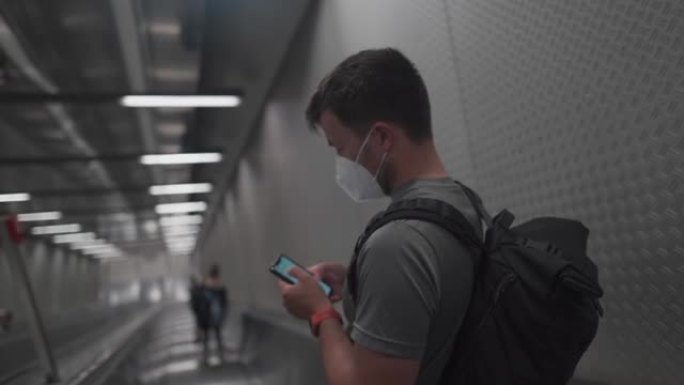 戴着防护口罩的乘客在地铁上下楼梯并使用手机。公共交通工具自动扶梯上的蒙面男子刷卡智能手机。地下移动楼