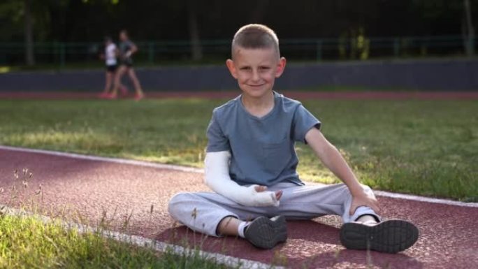 悲伤的断手小男孩在夏日坐在户外运动场上。9岁肢体骨折的孩子。骨折损伤。手臂上的石膏