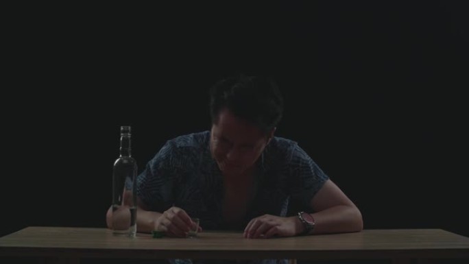 醉酒，沮丧的亚洲男子在喝酒前将伏特加酒倒入小玻璃杯中，并用拳头砸碎黑色背景的桌子