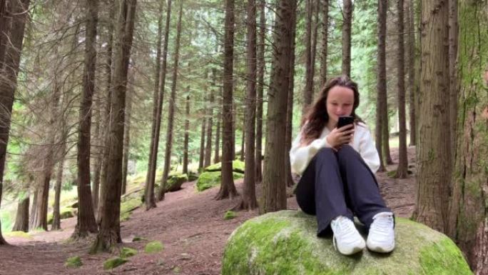 一个穿着白衬衫的女孩一个少年坐在森林里的大石头上她手里拿着黑色的iPhone 13她看着屏幕平静地举
