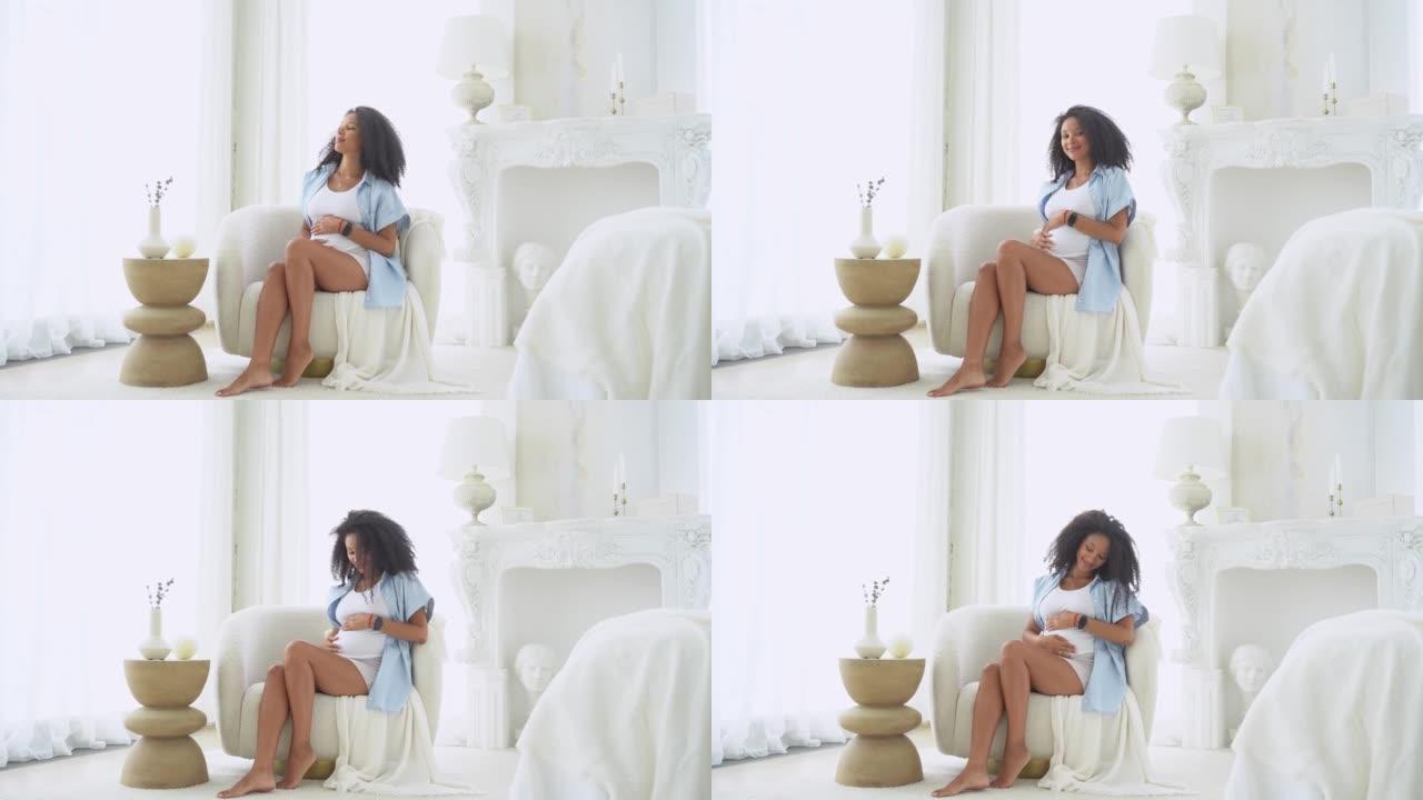 健康怀孕。黑色迷人的孕妇在白色卧室与大窗户坐在椅子上。美国黑人女性中孕期晚期妊娠，揉肚子。妈妈在等宝