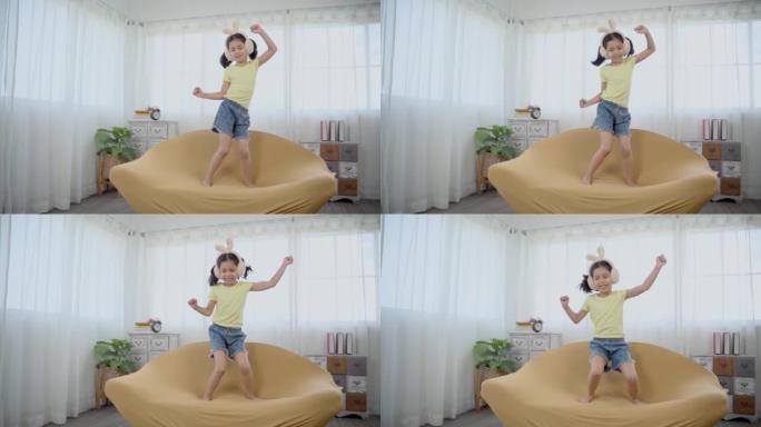 亚洲可爱的小女孩快乐地在沙发上跳舞和跳跃。有趣的孩子跳舞，用兔子耳机听音乐，享受音乐创造风格的舞蹈，