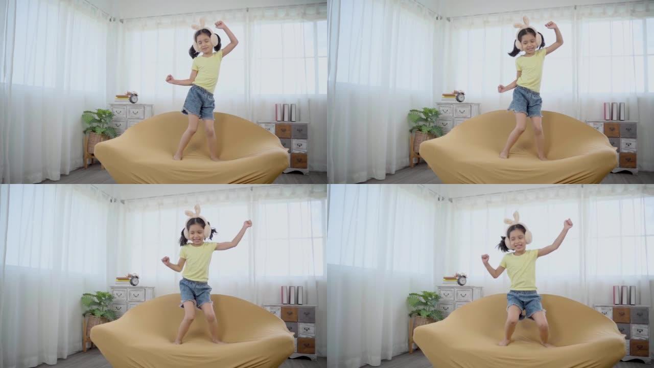 亚洲可爱的小女孩快乐地在沙发上跳舞和跳跃。有趣的孩子跳舞，用兔子耳机听音乐，享受音乐创造风格的舞蹈，