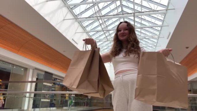 年轻的少女展示包裹她有了新的购买，她刚刚在一家大商店买东西，她在透明的屋顶上穿着白色衣服旋转和跳舞