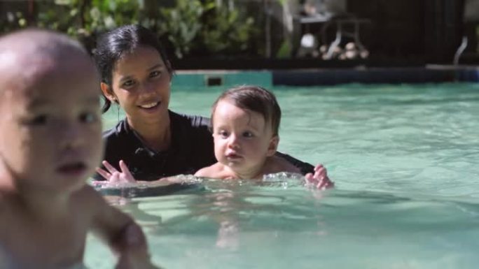 一位年轻的母亲双手抱着儿子在游泳池里的水中。一个10个月大的婴儿喜欢在温暖清澈的水中游泳。从小就教孩