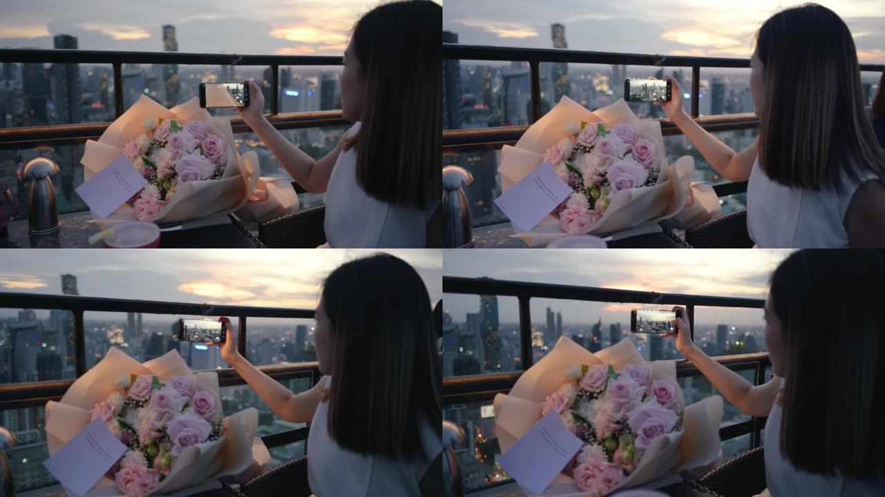 年轻的亚洲妇女手持玫瑰花束在日落时在城市豪华生活方式的屋顶庆祝生日在泰国曼谷吃晚餐户外微笑和快乐用餐