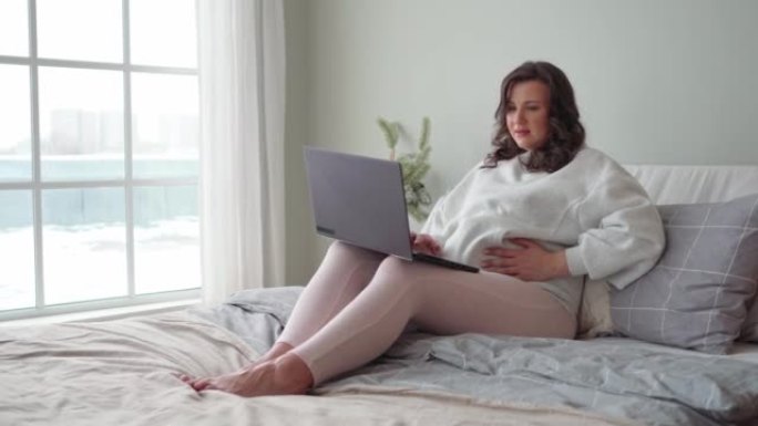 年轻的孕妇坐在床上，紧紧抓住并在笔记本电脑上工作