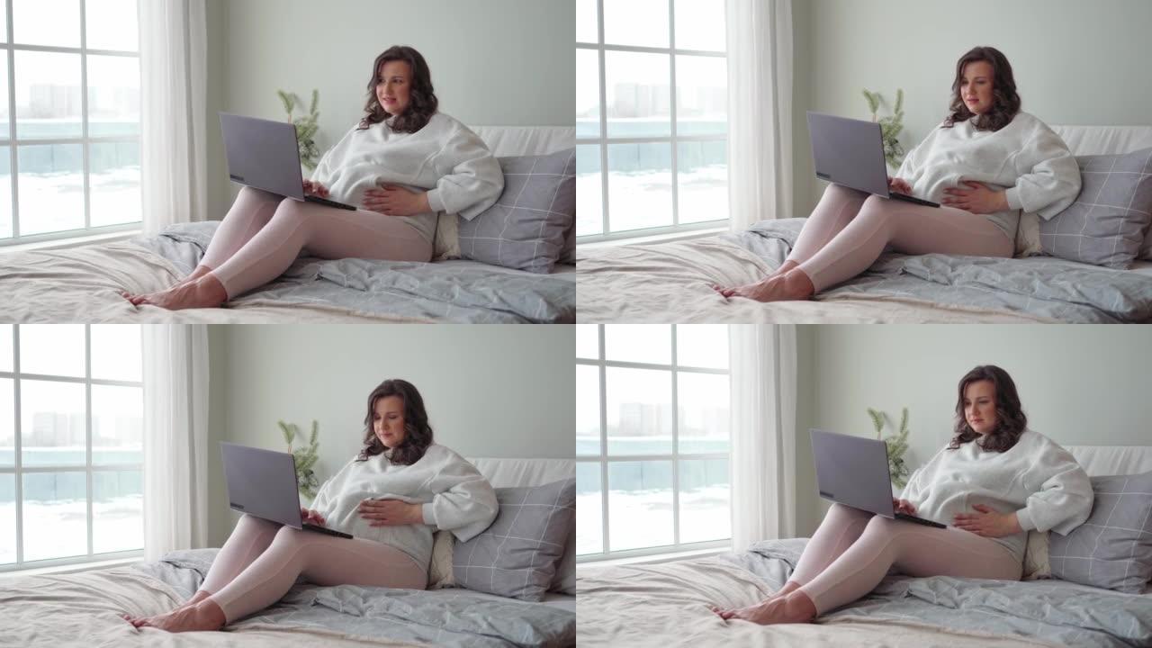 年轻的孕妇坐在床上，紧紧抓住并在笔记本电脑上工作