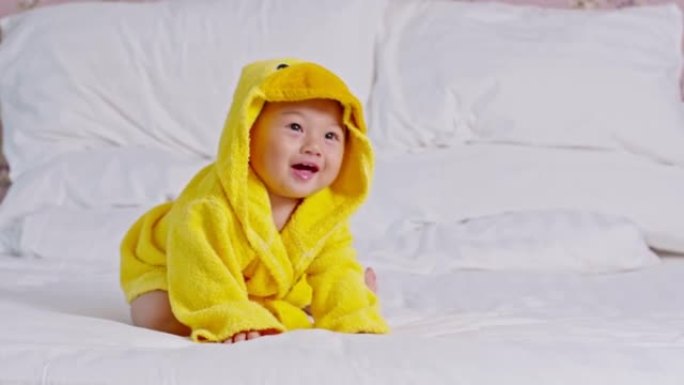 快乐的亚洲宝宝笑着穿着黄色的鸭子浴衣。洗澡时间。可爱的婴儿坐在床上，看着相机等待洗衣服和洗澡。婴儿护