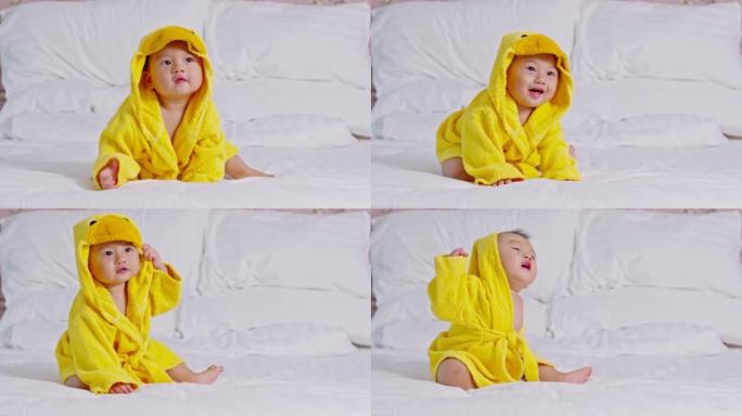 快乐的亚洲宝宝笑着穿着黄色的鸭子浴衣。洗澡时间。可爱的婴儿坐在床上，看着相机等待洗衣服和洗澡。婴儿护