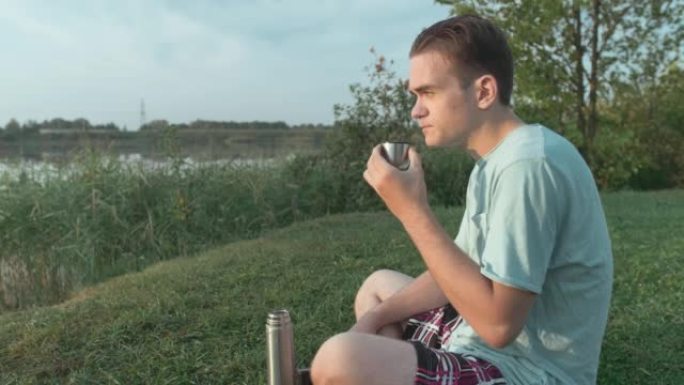 一个年轻人在湖后面的热水瓶里喝咖啡