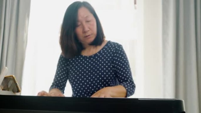 亚洲成年妈妈通过手机应用程序在家学习在窗户附近玩键盘乐器，生活方式概念。