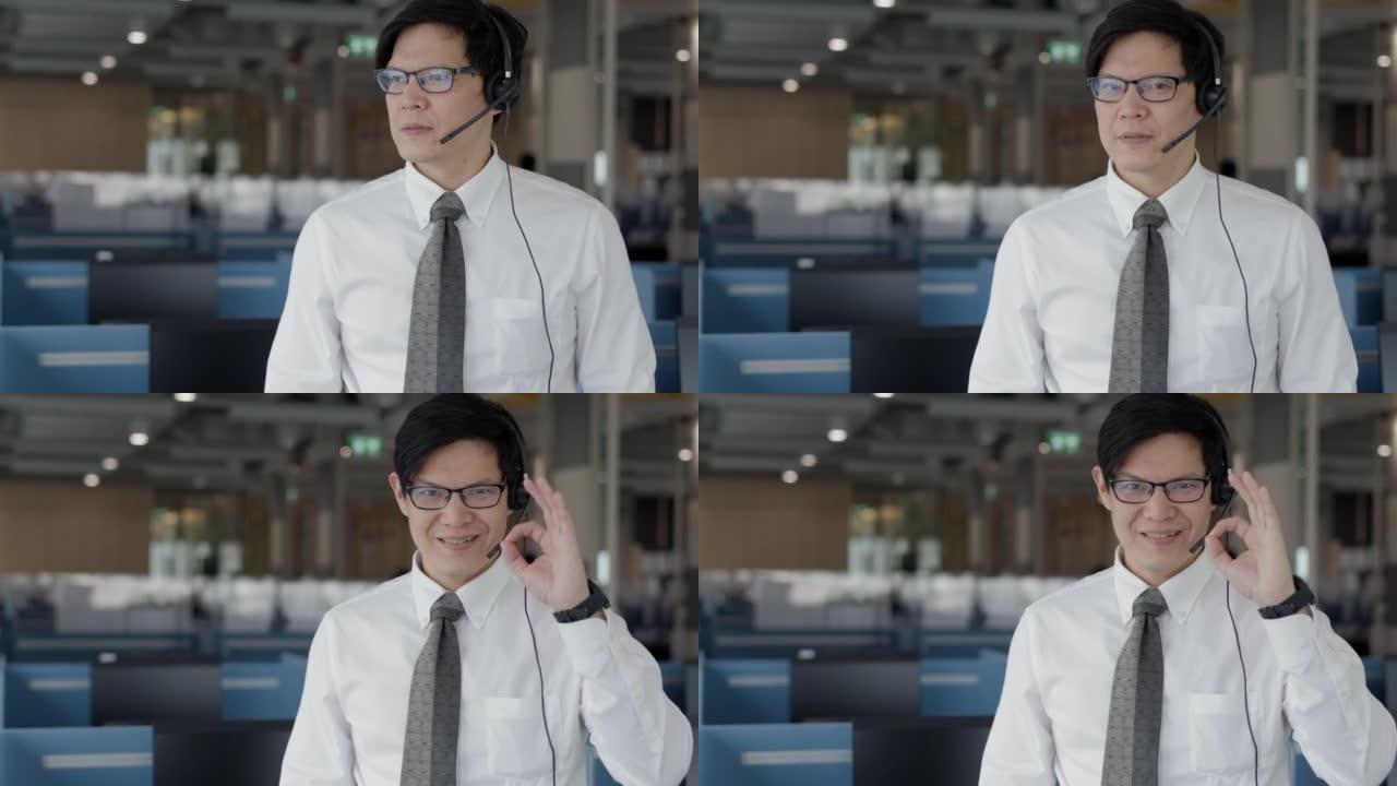 智能成功亚洲男性客户服务代表的肖像，带耳机，显示OK- sign，笑脸并站在呼叫中心房间。看着镜头。