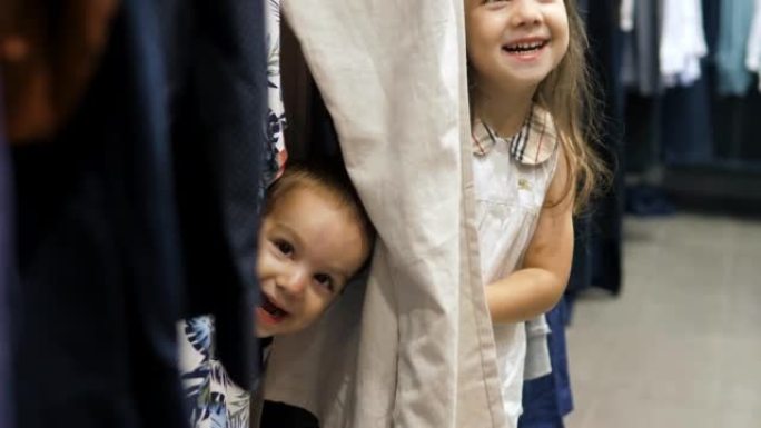 两个小孩在商店里玩耍，躲在东西里，在一家购物中心的服装店大声笑。孩子们玩得开心，在商店里玩得开心。生