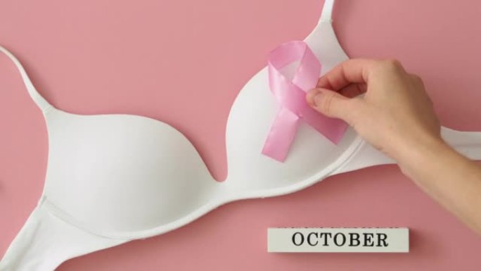 粉色背景乳腺癌宣传月。女人的手放一条粉红色的丝带-白色胸罩上的蝴蝶结。希望和恢复的象征。世界癌症日概