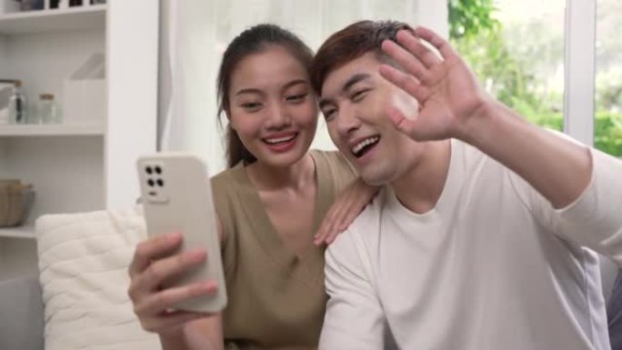 年轻的亚洲夫妇坐在一起工作，在家时使用智能手机在线会见朋友。