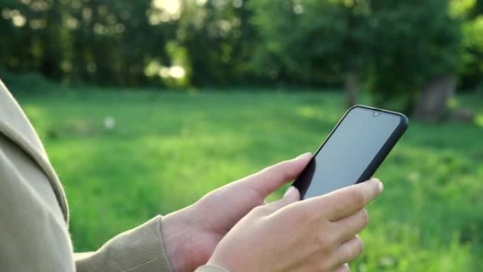 使用智能手机的女性双手特写镜头，自然背景。女人在户外日落时使用手机。无法识别的人触摸智能手机屏幕。不