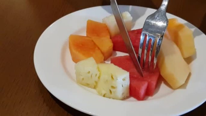 吃新鲜多汁的水果沙拉，木瓜，甜瓜，菠萝和西瓜配白盘健康早餐