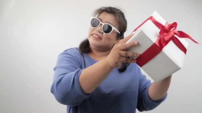 胖胖的成年女性戴着墨镜拿着礼品盒，在灰色背景上跳舞，假日生活方式概念。