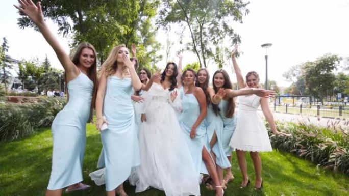 美丽的新娘和她漂亮的新娘穿着同样的淡蓝色连衣裙，在公园里欢呼挥手。穿着白色礼服派对的长而通风的新娘面