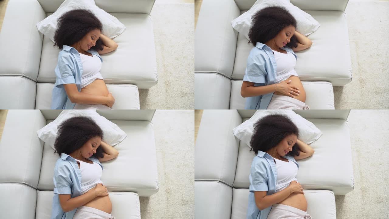 怀孕的黑人美女躺在家里的沙发上，摸着肚子，梦见未来。放松和有意识怀孕的健康概念。