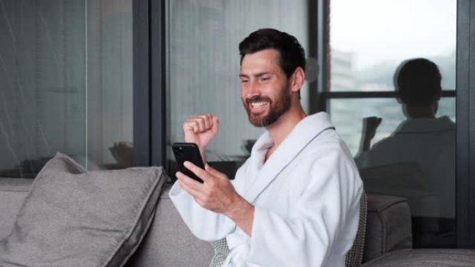 穿着浴袍庆祝成功赢家的兴奋快乐商人在酒店公寓看手机。特写快乐的人在电话中以慢动作阅读好消息。惊讶的幸
